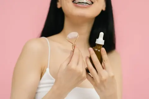 Uśmiechnięta osoba z aparatem ortodontycznym trzyma roller do twarzy i buteleczkę z serum. Idealne połączenie: retinol i kwas hialuronowy dla pięknej cery.