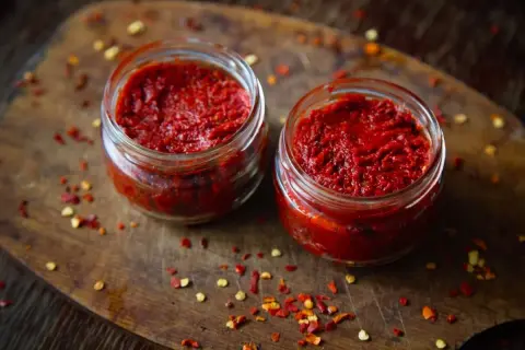 Dwa słoiczki z adżiką, idealną do doprawienia dań. Na drewnianej desce rozsypane płatki chili.