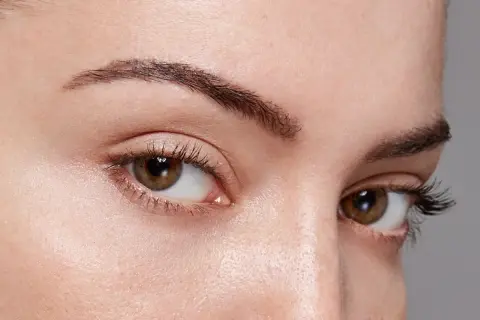 Gros plan sur des yeux marron avec des cils longs. Le sourcil gauche est légèrement plus fin, montrant comment dessiner ses sourcils pour un look naturel.