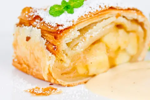 Ein Stück Apfelstrudel mit Puderzucker und Minze, serviert mit Vanillesauce.