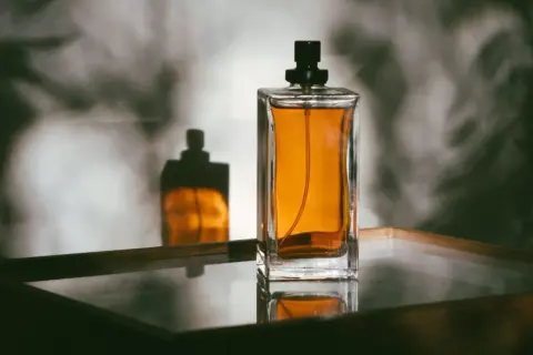 Szklany flakon perfum z bursztynowym płynem i czarną nakrętką, odbijający się w lustrzanej powierzchni.