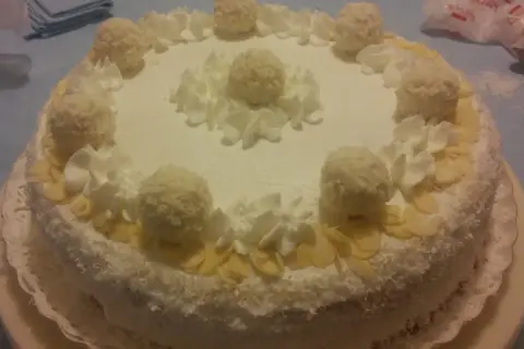 Torta Raffaello ricoperta di cocco, decorata con panna montata e praline, un'esplosione di dolcezza.