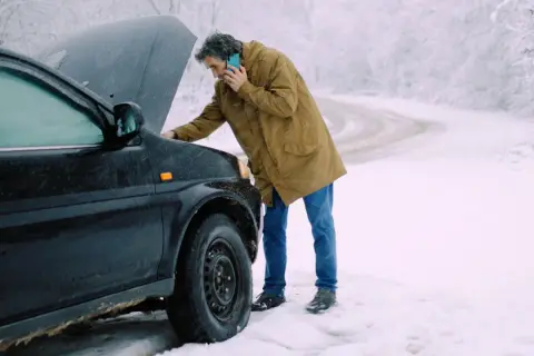 Man med telefon vid en bil med öppen motorhuv. Kallstart bil i snöväder.