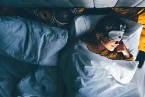 Ein Mädchen mit Hasen-Schlafmaske liegt im Bett und scheint schnell einschlafen zu wollen.