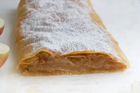 Ein köstlicher Apfelstrudel mit fertigem Strudelteig, bestäubt mit Puderzucker. Daneben liegen frische Apfelstücke.