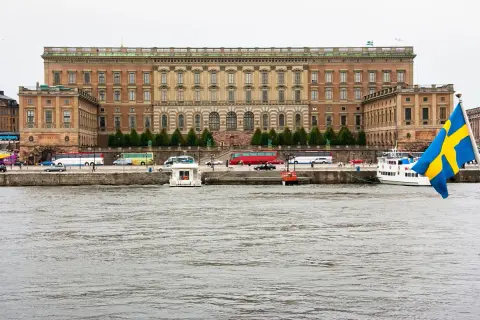Stor byggnad vid vattnet, troligen kuninkaanlinna tukholma, med bussar och båtar. Svenska flaggan vajar.