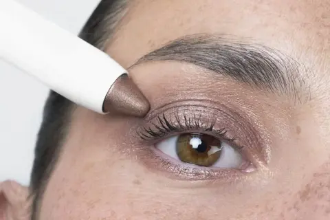 Application d'un fard à paupières bronze sur des yeux marrons. Un bâtonnet blanc dépose la couleur sur la paupière.