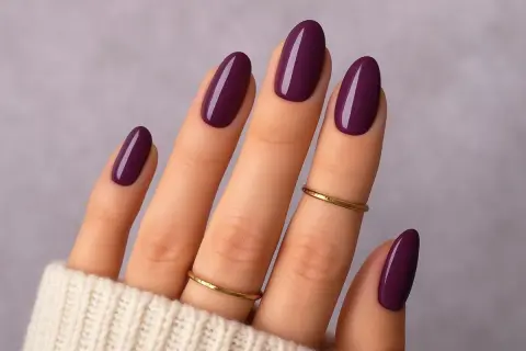 Main d'une femme aux ongles vernis d'un prune profond, une parfaite idée ongle janvier. Deux fines bagues dorées ornent les doigts.