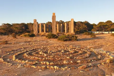 Steine bilden ein Labyrinth im Freien, umgeben von Pinien. Im Hintergrund ragen steinerne Säulen auf, die an Zeit und Raum auf Ibiza erinnern.