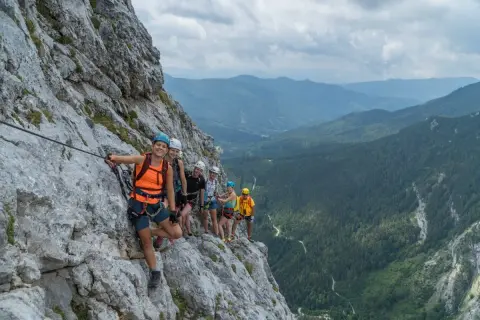 Grupa uśmiechniętych ludzi na via ferrata w Dolomitach, zabezpieczona linami, podziwia malownicze widoki górskiego krajobrazu.