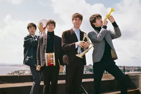 The Beatles grają na instrumentach, tworząc piosenki. George gra na trąbce, John na harfie, Paul na tamburynie, a Ringo ma bębenek na szyi.