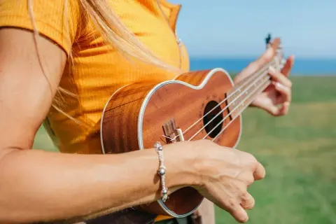 Kobieta w żółtej koszulce gra na ukulele na tle zielonej łąki i morza.
