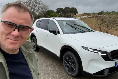 Mężczyzna w okularach pozuje do selfie obok białej Mazdy CX-5 na tle winnicy.
