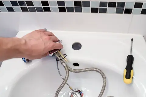 Hand montiert einen Wasserhahn, um das Waschbecken zu reparieren. Ein Schraubenzieher liegt bereit.