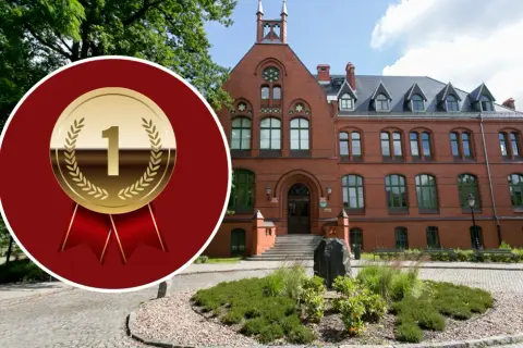 Złoty medal z cyfrą 1 na tle ceglanego budynku i zieleni.