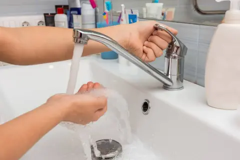 Mano ajustando el grifo para aumentar la presión del agua mientras se lava las manos en un lavabo blanco.
