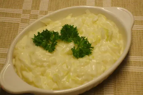 En vit oval skål med krämig stuvad vitkål, toppad med färsk persilja. Serveras på en rutig duk.