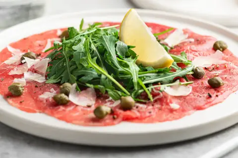 En tallrik med tunnskivad carpaccio di manzo, toppad med rucola, kapris, parmesan och en citronklyfta.