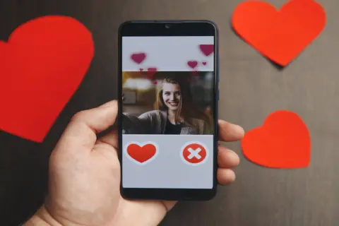 Dłoń trzyma smartfon z ekranem aplikacji randkowej. Widoczna uśmiechnięta dziewczyna i przyciski serca i krzyżyka. Wokół czerwone serca.