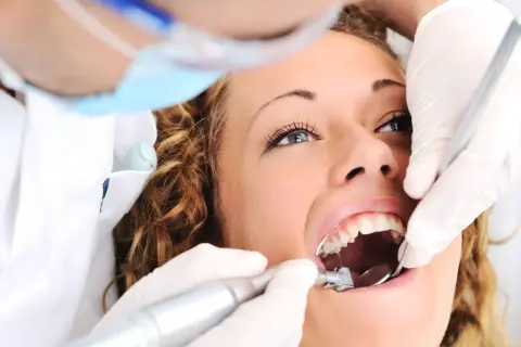 Kobieta u dentysty, zastanawia się ile kosztuje plomba prywatnie. Dentysta w rękawiczkach używa wiertła i lusterka.