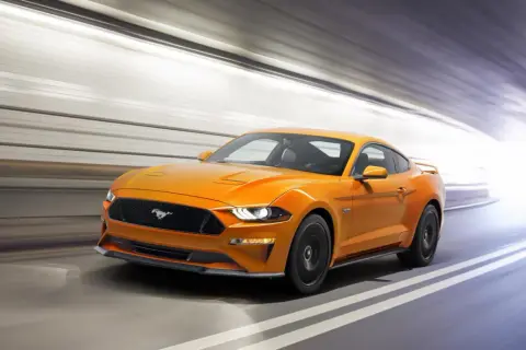 Un Mustang naranja acelera en un túnel, sus potentes sonidos de coches deportivos resonando.