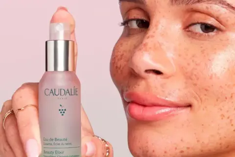 Una mano sostiene el Agua de Belleza Caudalie, mientras una mujer con pecas sonríe. Opiniones positivas sobre este elixir para una piel radiante.