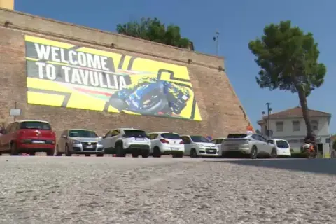 Benvenuti a Tavullia, la terra di Valentino Rossi! Un grande striscione giallo celebra il campione, con auto parcheggiate sotto.