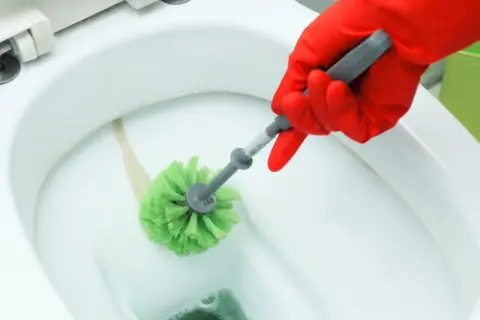 Mano con guanto rosso pulisce il WC con uno scopino verde per togliere il calcare.