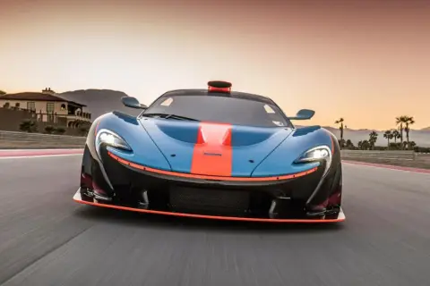 El McLaren P1, azul con franjas naranjas, acelera en una pista al atardecer, con palmeras y montañas de fondo.