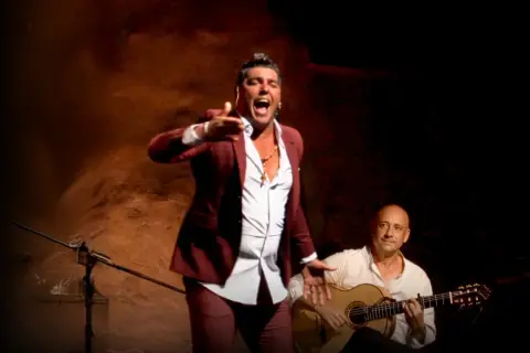 Un hombre con traje granate canta con pasión, su voz flamenco resuena mientras un guitarrista lo acompaña.