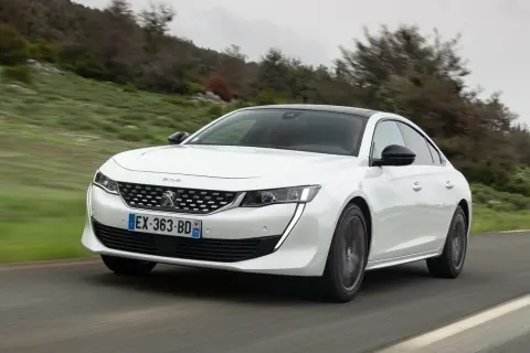 Biały Peugeot 508 jedzie po krętej drodze wśród zielonych wzgórz.