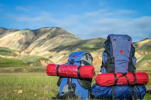 Dos mochilas de montaña listas para la aventura, con sacos de dormir rojos sujetos. El paisaje montañoso de fondo inspira a saber como llevar una mochila de montaña.