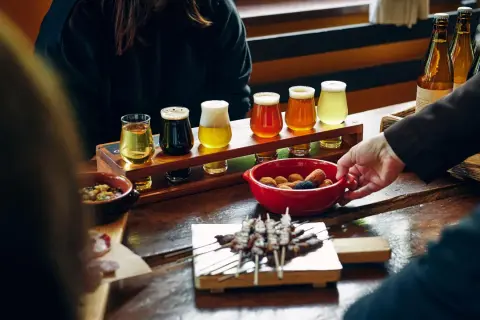 Degustazione di birre pregiate: un assortimento di diverse birre in bicchierini, accompagnate da stuzzichini e spiedini su un tavolo di legno.