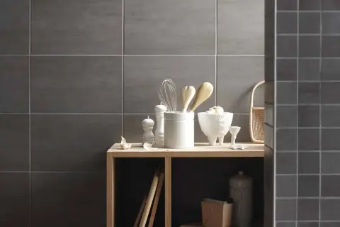 Sur un mur en carrelage gris, des ustensiles de cuisine et des objets décoratifs sont disposés sur une étagère en bois. La faïence du mur de droite est plus petite.