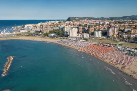 Vista aerea della Pesaro strand, con ombrelloni colorati, mare azzurro e la città sullo sfondo.