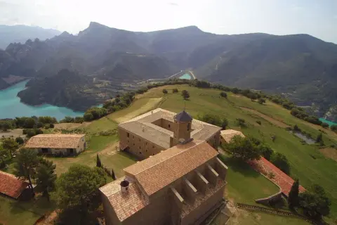 Edificio histórico en la Serra de Busa, con un lago turquesa y montañas al fondo.