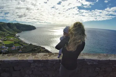 Widok na wybrzeże z małą wioską i oceanem. Kobieta trzyma dziecko, podziwiając krajobraz. Azory czy warto? Tak!