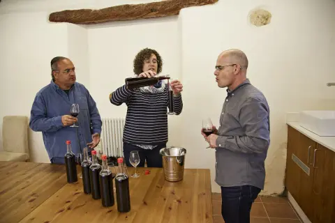 Weinprobe: Eine Frau gießt Wein ein, während zwei Männer zuschauen. Flaschen und Gläser stehen bereit für die assemblage wein.