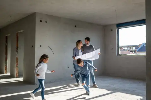 Rodzina ogląda plany w mieszkaniu w stanie deweloperskim. Dzieci biegają po pustym pokoju, rodzice analizują projekt.