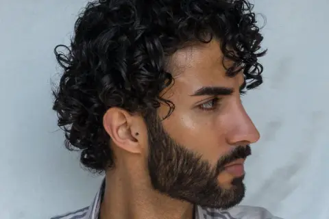Hombre con un moderno peinado chino, cabello rizado oscuro y barba cuidada.