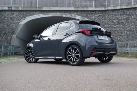 Szary Toyota Yaris GR Sport z czarnym spojlerem i dyfuzorem, zaparkowany przed tunelem z kamiennym murem.
