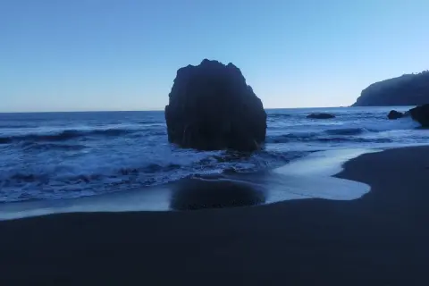 Olas rompen contra una gran roca en la playa los patos. El cielo azul claro se funde con el mar.