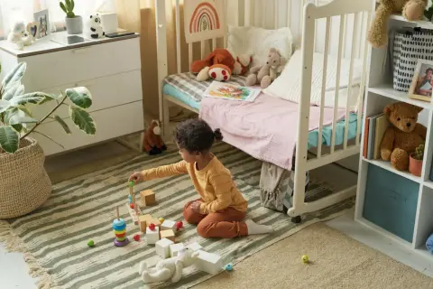 Ein kleines Kind spielt mit Holzbausteinen in einem gemütlichen, quadratischen Kinderzimmer. Spielzeug und ein Bett laden zum Verweilen ein.