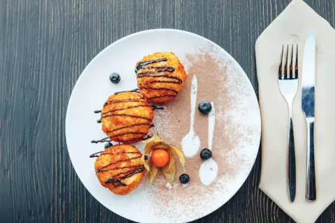 Drei goldbraune, knusprige Kugeln, beträufelt mit Schokoladensauce, liegen auf einem Teller mit Kakaopulver und Beeren. Ein süßer Genuss, der an Eis erinnert, aber als knusprige Leckerei serviert wird.