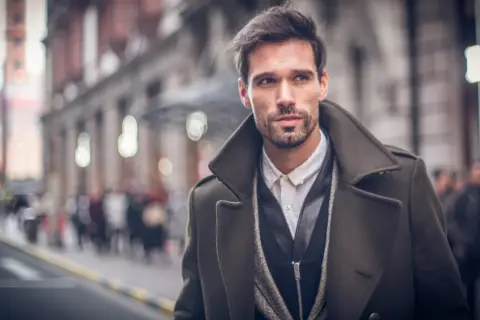 Ein Mann mit Bart trägt eine elegante, olivgrüne Jacke zum Anzug. Er blickt nach rechts, während er durch eine belebte Stadt geht.