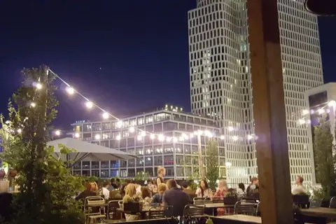 Abendstimmung auf einer Dachterrasse in Berlin. Lichterketten beleuchten Tische, an denen Gäste sitzen. Im Hintergrund ragen moderne Gebäude auf, die an den Spreegold Bikini erinnern.