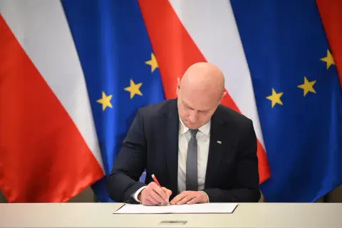 Łysy mężczyzna w garniturze podpisuje dokument na tle flag Polski i UE. Spodziewaj się niespodziewanego, bo nigdy nie wiadomo, co przyniesie przyszłość.