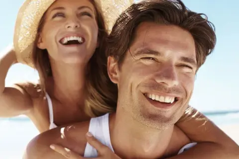 Glückliches Paar am Strand, die Frau lacht und trägt einen Hut. Selbst im Schatten wird man braun, wenn man die Sonne genießt.