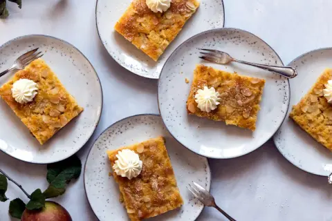 Saftiger Apfelkuchen vom Blech, garniert mit Sahnetuffs und Mandelblättchen, auf Tellern angerichtet.