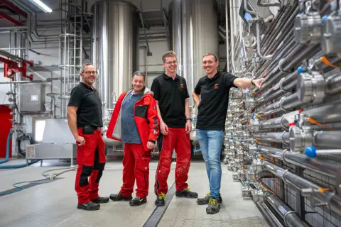 Vier Männer in Arbeitskleidung stehen vor großen Edelstahltanks und Rohrleitungen. Sie sind Teil einer Ausbildung zum Brauer und blicken stolz in die Kamera.